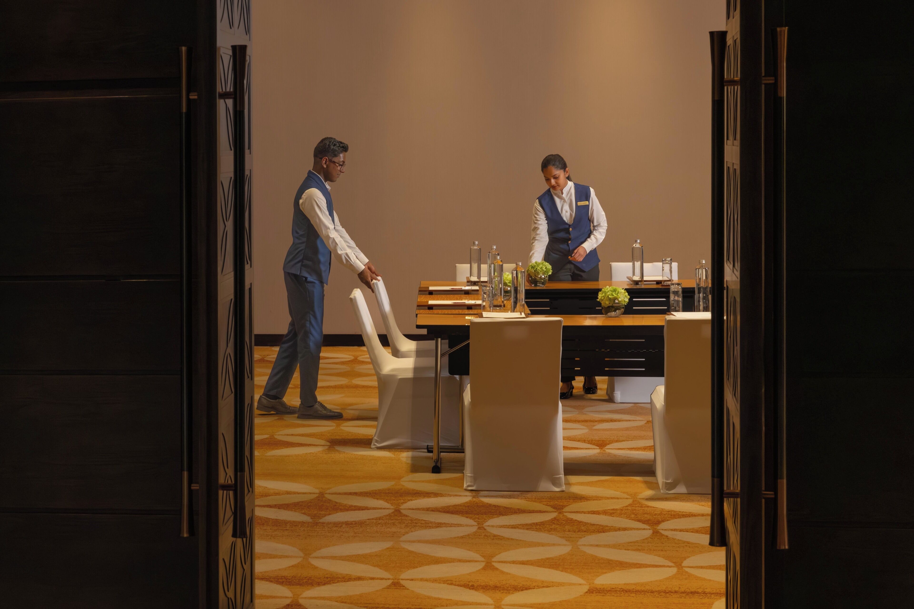 A hotel room with a wooden table set for two, a white chair, and a patterned carpet.