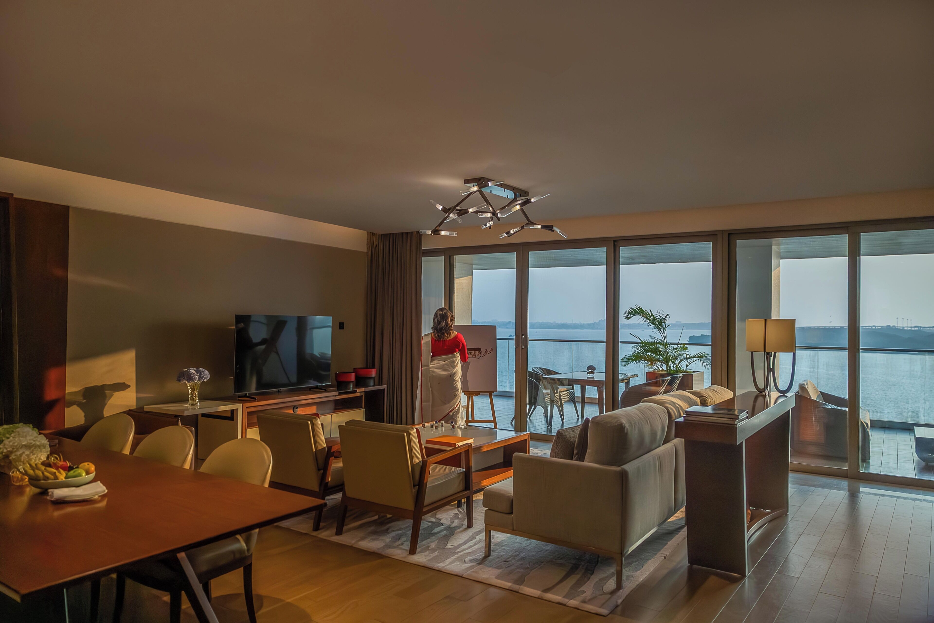 A modern hotel room with a large TV, dining area, sofa, and a view of the sea.