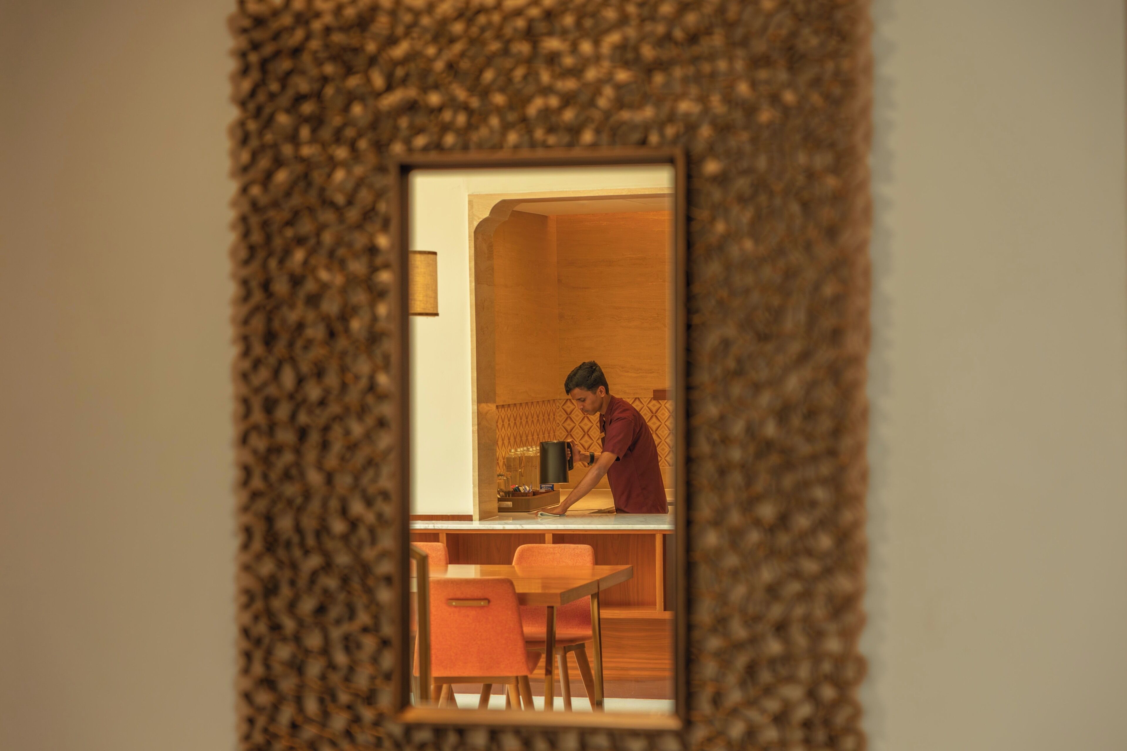 A kitchen with a person cooking, a wooden cabinet, and a small dining area with a chair.