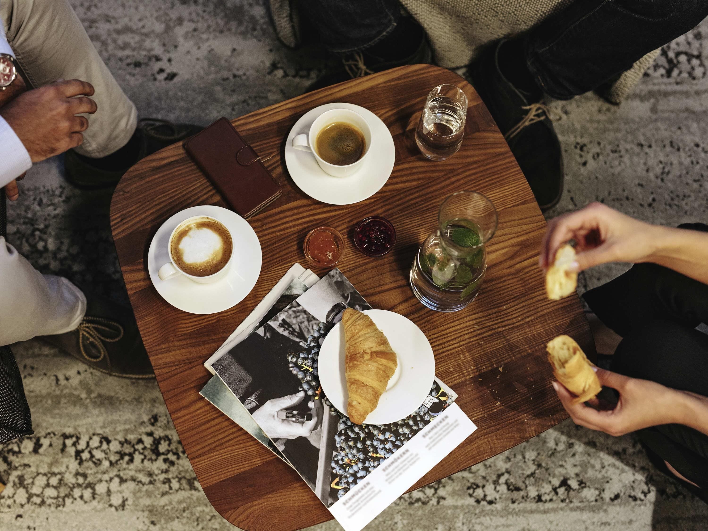 Ein Tisch mit einem Croissant, einem Glas Wasser, einem Glas Saft, einer Tasse Kaffee und einer Zeitschrift.