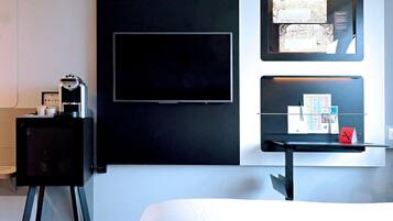 Habitación de hotel con una televisión de pantalla plana montada en la pared, una cafetera pequeña, un escritorio con una bandeja de objetos y un tablón de anuncios con papeles y un letrero rojo.
