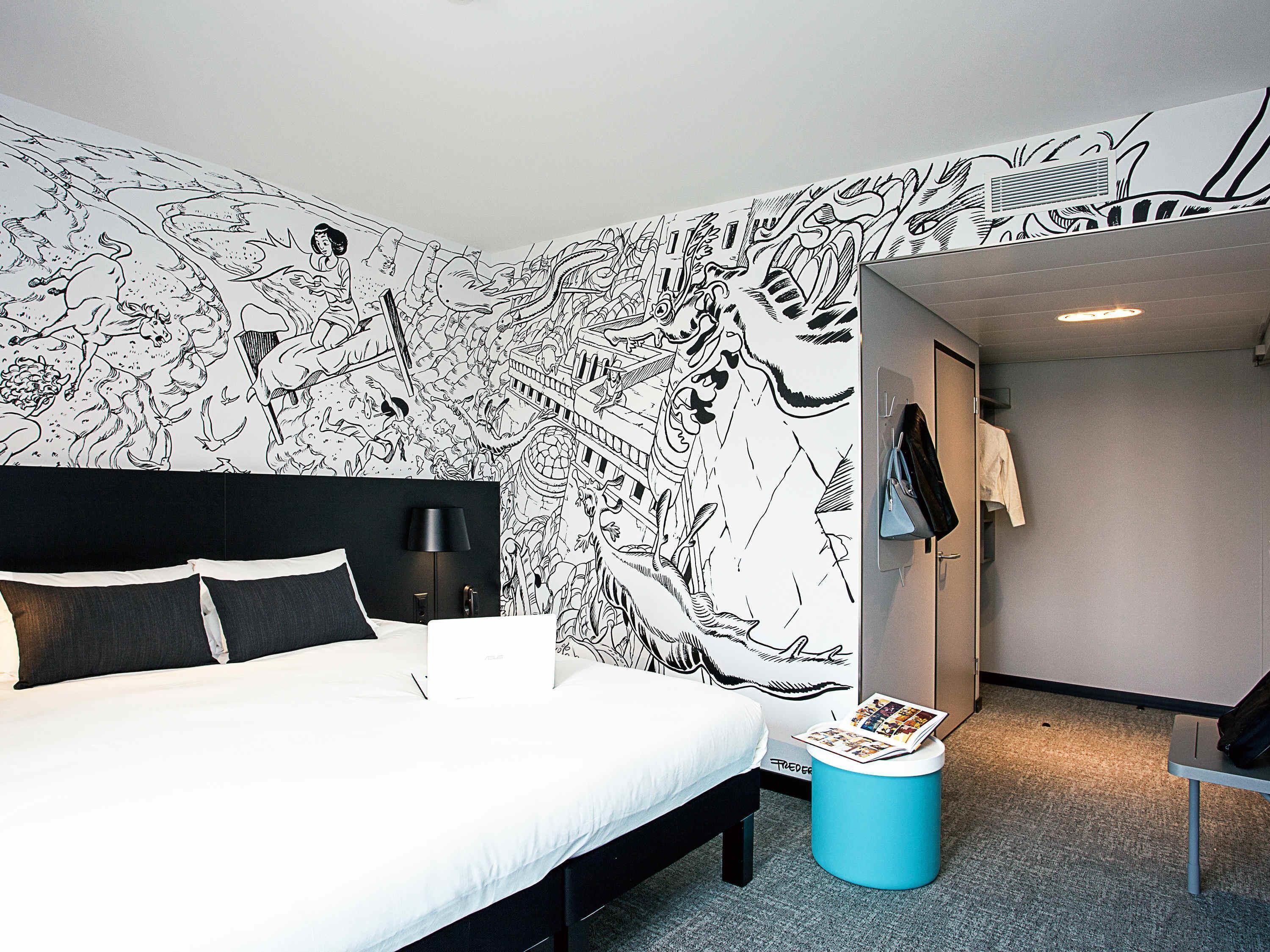 A hotel room with a large black and white mural on the wall, a bed with black headboard, a blue ottoman, and a small table with a magazine.