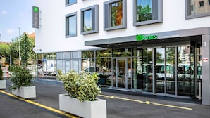 Um hotel moderno com fachada de vidro, uma placa verde indicando que pertence à rede ibis styles, e o número 3 no edifício.