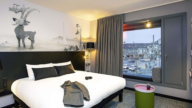 Quarto de hotel com cama, vista da cidade, um quadro na parede e uma lixeira verde.