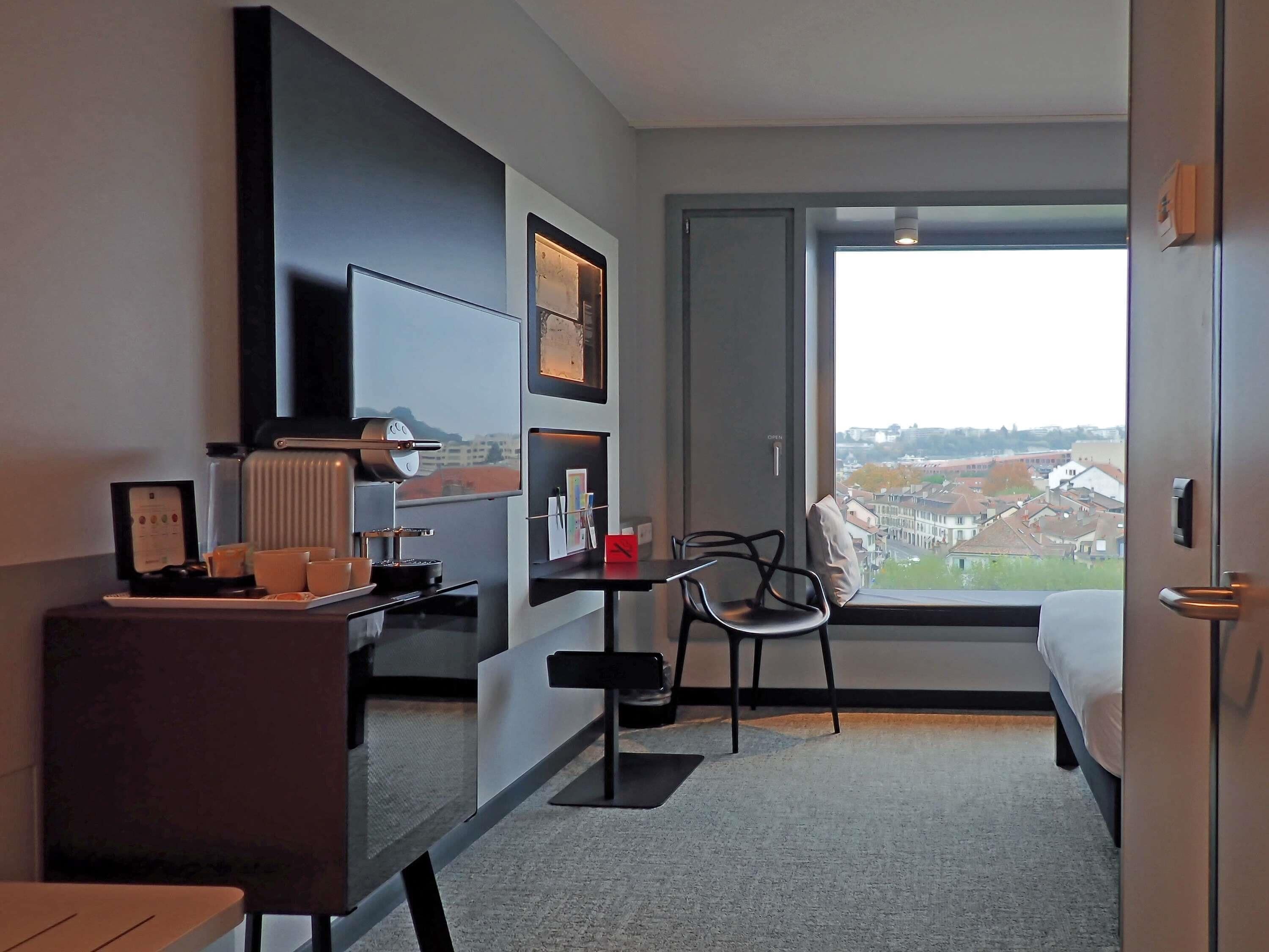 A hotel room with a desk, chair, bed, and a large window offering a city view.