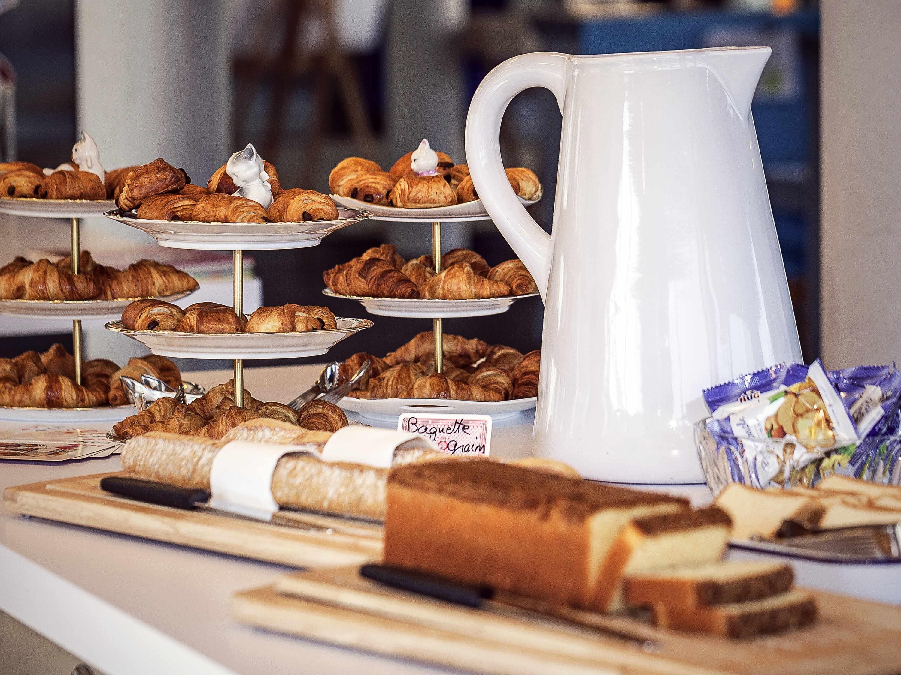 A display of pastries and bread, including croissants and baguettes, with a white pitcher and a sign indicating 'Baguette Pain'.
