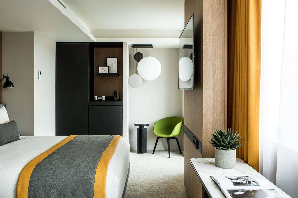 A modern hotel room with a bed, a desk with a plant, a mirror, and a green chair.