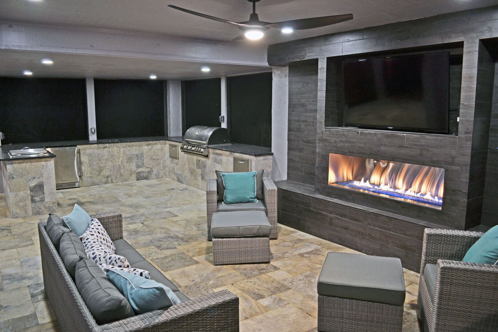 An outdoor patio with a stone floor, wicker furniture, a built-in grill, and a flat-screen TV above a fireplace.