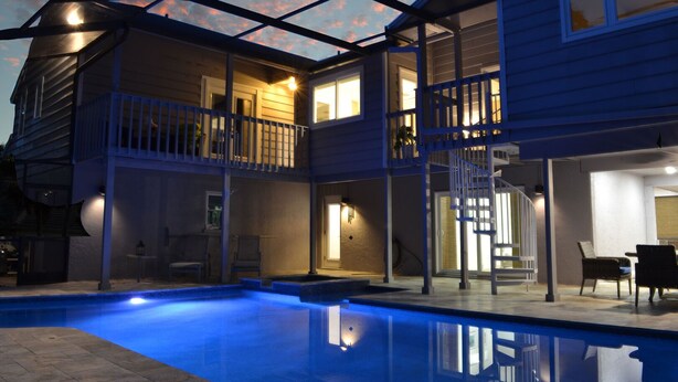Una casa de dos pisos con piscina, mobiliario de exterior y un patio cubierto.