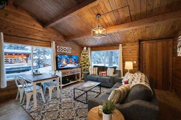 A cozy cabin interior with a wooden ceiling, a TV on a wooden stand, a Christmas tree, a sofa, and a dining area with chairs and a table. - Near Lake Wenatchee|Fence|Hot Tub|WIFI|Dogs|Sleep4 (Leavenworth)