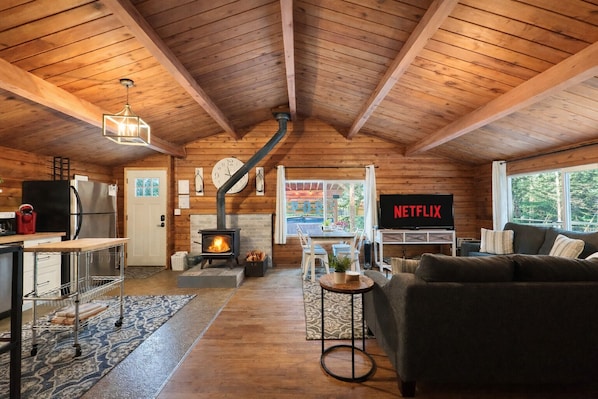 A cozy cabin interior with a fireplace, a kitchen area, a living room with a sofa, and a TV displaying 'NETFLIX'. - Near Lake Wenatchee|Fence|Hot Tub|WIFI|Dogs|Sleep4 (Leavenworth)