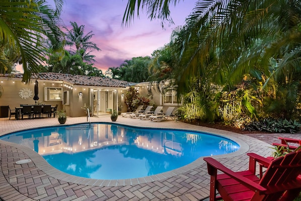 A swimming pool with lounge chairs, a dining area, and a building with outdoor lighting.