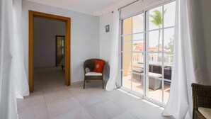 A room with a large sliding glass door leading to an outdoor area, a wicker chair with a red cushion, and a wooden door frame. - Estrella Del Mar H6_200Mbps_BBQ_Pool_Walk to Beach (Bavaro-Punta Cana)