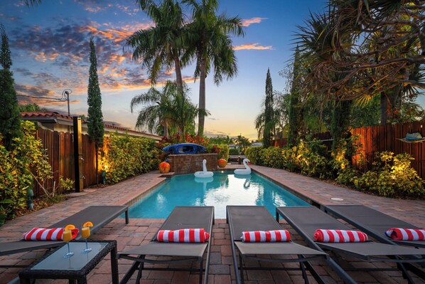 A swimming pool with lounge chairs and swan floaties, surrounded by palm trees and a wooden fence.