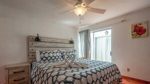 Une chambre à coucher comprenant un lit, une table de chevet, un ventilateur de plafond, une fenêtre avec des rideaux et un tableau mural.