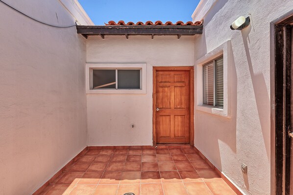 Un estrecho pasillo exterior con suelo de baldosas, una puerta de madera y una ventana con persianas.