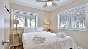 Une chambre à coucher avec un lit, deux lampes de chevet, un ventilateur de plafond et des fenêtres donnant sur la forêt.