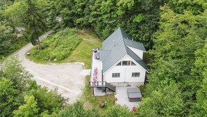Une maison avec un toit en pente, entourée d’une forêt dense, avec une allée en gravier et un petit espace patio.