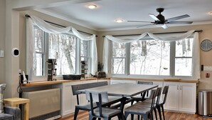 Une cuisine avec une table à manger, des chaises, un ventilateur de plafond et de grandes fenêtres.