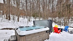 Un bain à remous installé à l’extérieur, avec en arrière-plan une forêt enneigée, et entouré de fauteuils Adirondack.