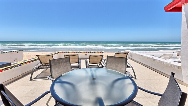 Een terras aan het strand met een ronde tafel en vier stoelen, met uitzicht op de oceaan.