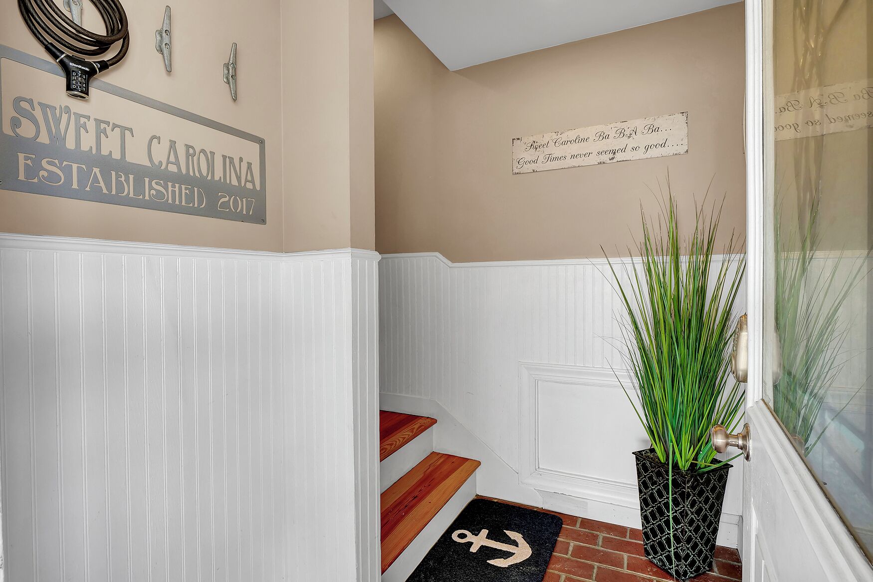 A hallway with white wainscoting, a sign reading "Sweet Carolina Established 2017", a decorative sign with a quote, and a potted plant with tall green grass.