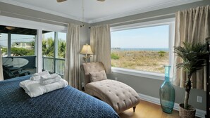 Ein Schlafzimmer mit Bett, Sessel, Lampe, einem großen Fenster mit Blick auf den Strand und einem Deckenventilator.