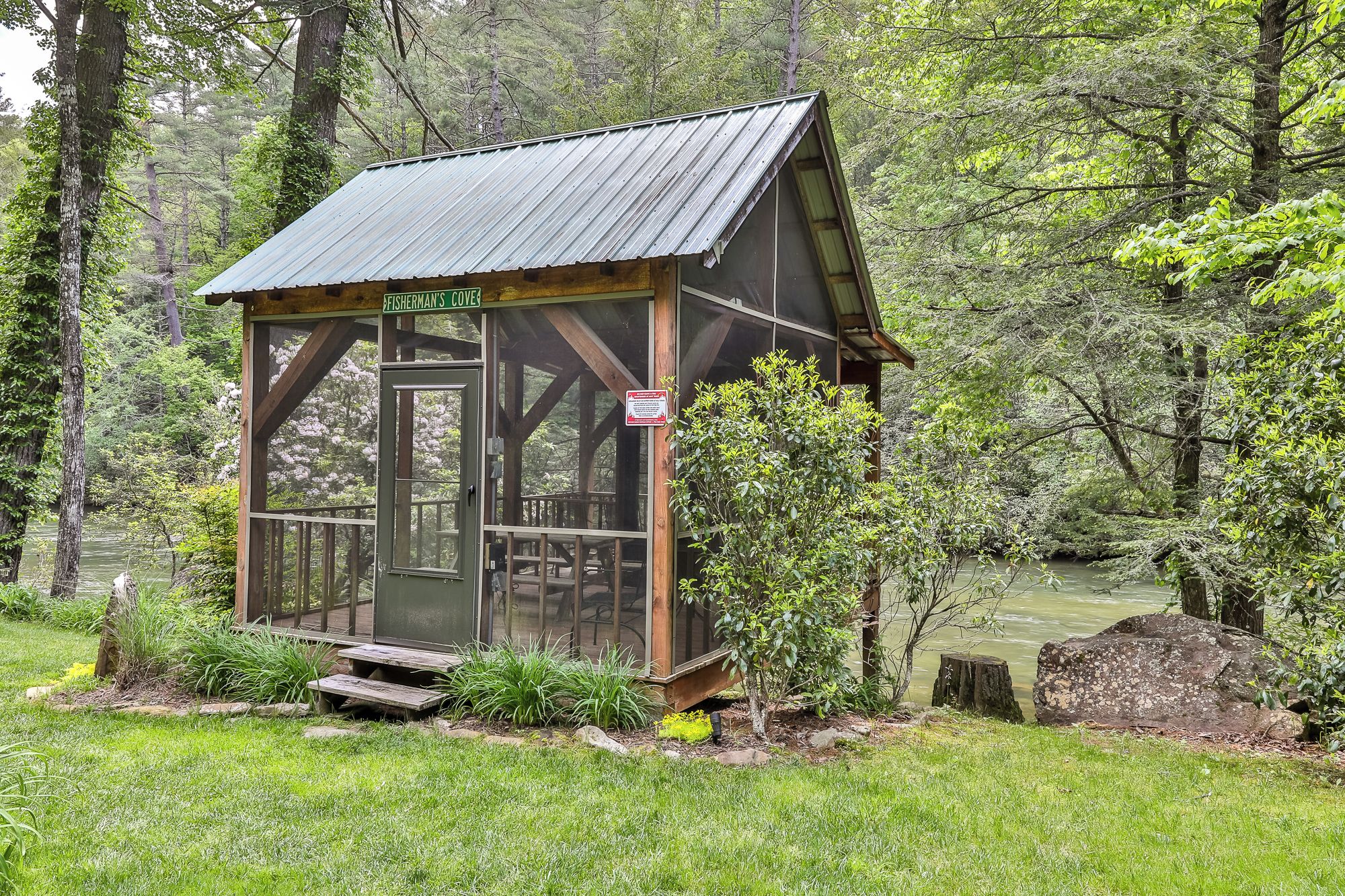 A small wooden cabin with a metal roof, surrounded by lush greenery and a body of water.