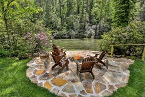 A stone patio with wooden chairs and a fire pit, surrounded by lush greenery and a view of a river. - PICK'S RIVERSIDE RETREAT - 2 Bedroom plus Loft Riverside Retreat on the Upper Toccoa River (Blue Ridge)