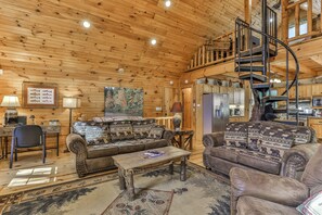 L’intérieur d’une cabane en rondins, avec un plafond en bois, un coin repas, une cuisine et un escalier en colimaçon.