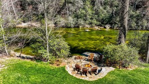 A stone fire pit with chairs and a fire, surrounded by a grassy area and trees, with a pond in the background.