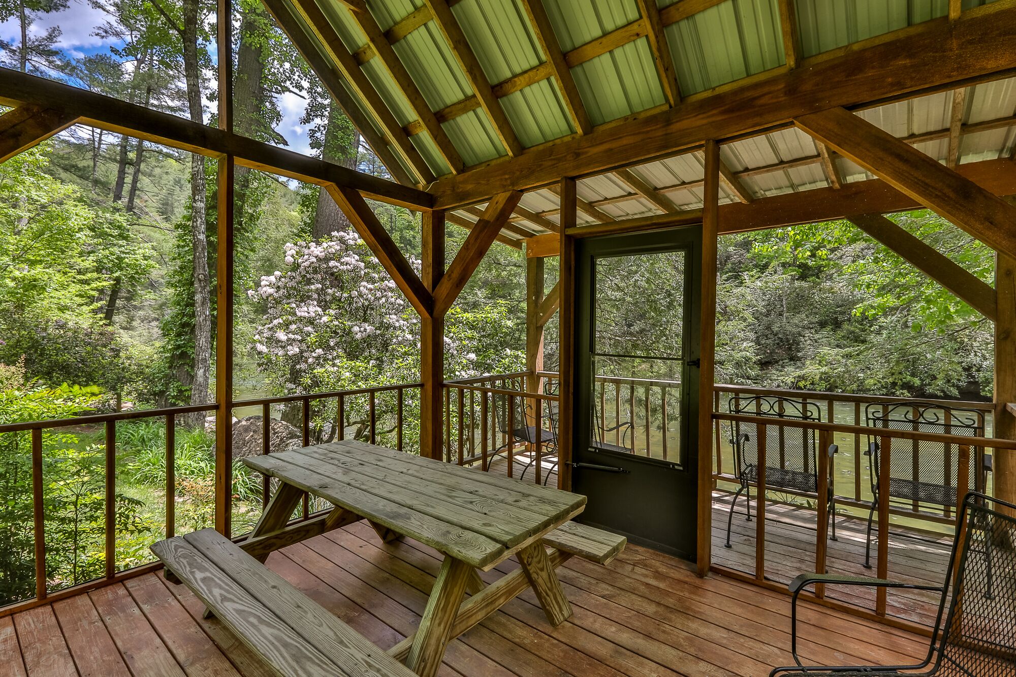 A covered wooden deck with a picnic table and benches, surrounded by metal railings, overlooking a forested area.