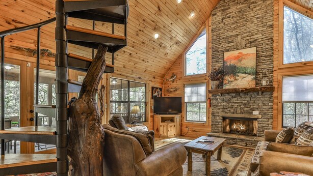 A spacious living room with a stone fireplace, wooden ceiling, and large windows.