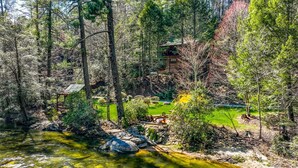A cabin surrounded by dense forest, a small pond, and a fire pit area.