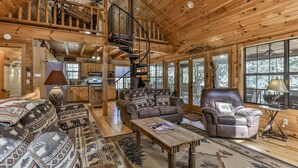 A spacious log cabin interior with a central living area featuring a sofa, armchairs, and a coffee table. A spiral staircase leads to an upper level with a kitchen and dining area. Large windows provide views of the surrounding forest.
