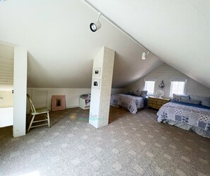 A room with a sloped ceiling, two beds with patterned bedding, a white pillar, and a small table with a fan.