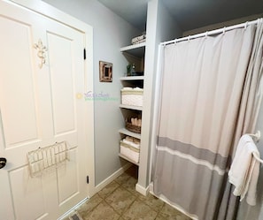 A bathroom with a white door, a shower curtain, and shelves with towels and toiletries.