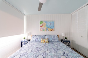 Une chambre à coucher comprenant un lit, deux tables de chevet, un ventilateur de plafond, un tableau accroché au mur et une armoire.