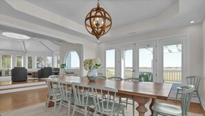 A spacious dining area with a wooden table and white chairs, large windows, and a view of the outdoors. - RARE Oceanside 5BR Waterfront Home w/ pvt Dock! (Wrightsville Beach)