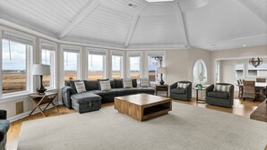 A spacious living room with a large sectional sofa, a wooden coffee table, and a view of the outdoors through large windows. - RARE Oceanside 5BR Waterfront Home w/ pvt Dock! (Wrightsville Beach)
