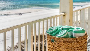 A balcony with a wicker basket containing rolled towels, overlooking a beach with a clear view of the ocean.