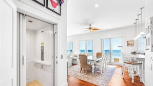 A spacious living area with a wooden floor, a dining table with chairs, a ceiling fan, and large windows offering a view of the ocean.