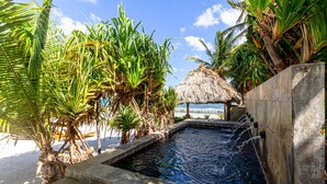 Ein Schwimmbecken mit Holzterrasse, umgeben von tropischer Bepflanzung und einer Struktur mit Reetdach.