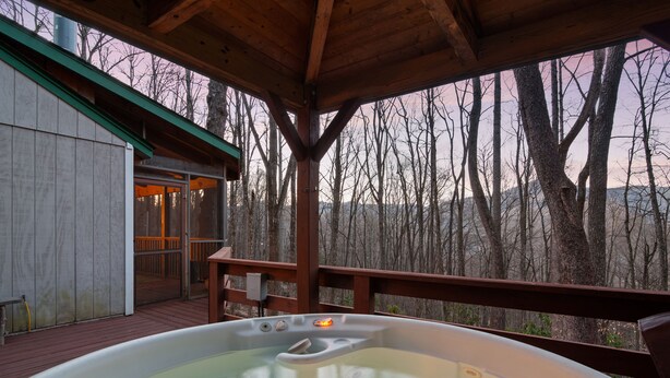 A hot tub in a wooden deck area with a view of a forested landscape. - 2 Master Suites - Hot Tub - Porch - Fire Pit - Wi-Fi - Clean - Cozy - 5 Stars! (Maggie Valley)