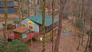 A cabin with a green roof, a deck, and a small outdoor seating area. - 2 Master Suites - Hot Tub - Porch - Fire Pit - Wi-Fi - Clean - Cozy - 5 Stars! (Maggie Valley)