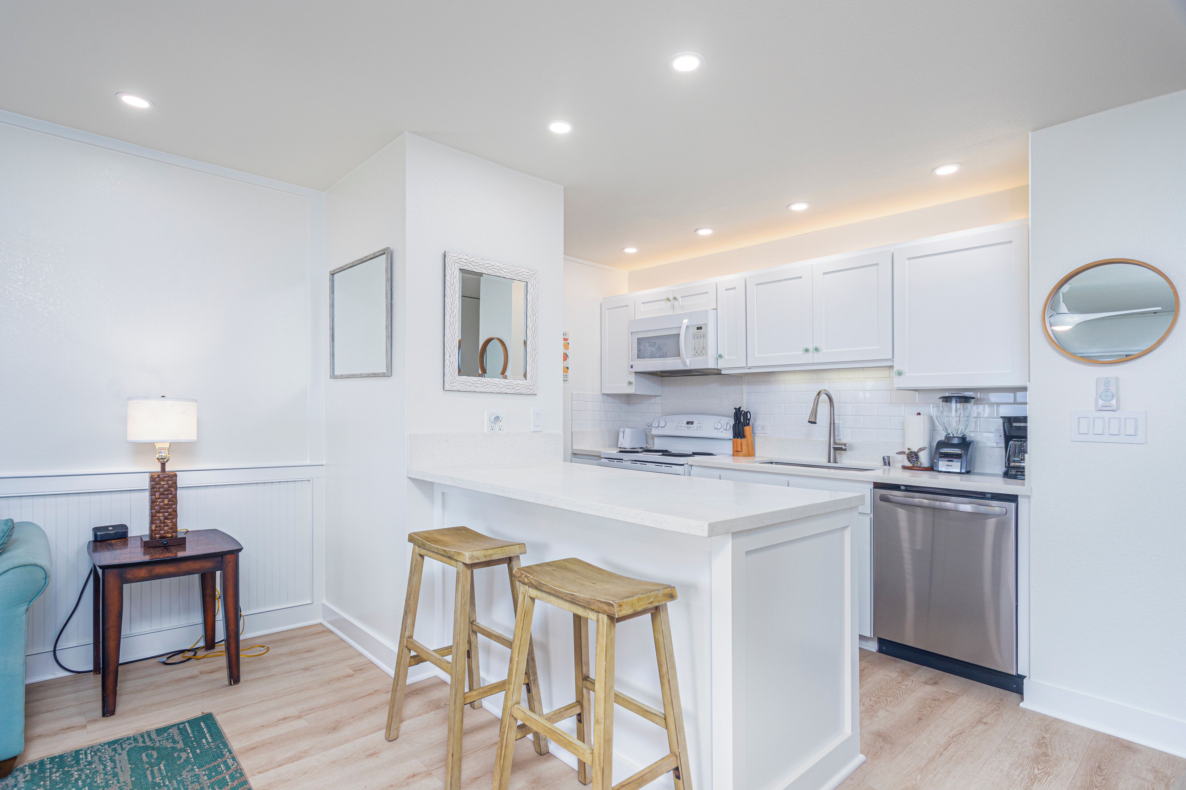 A modern kitchen with white cabinetry, a central island with stools, a microwave, a dishwasher, and a small table with a lamp.