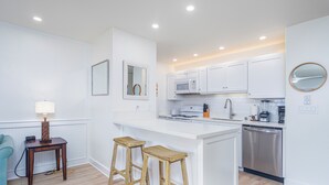 Une cuisine moderne avec des meubles blancs, un îlot central doté de tabourets, un four à micro-ondes, un lave-vaisselle et une petite table avec une lampe.