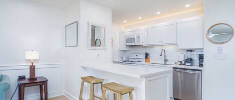 A modern kitchen with white cabinetry, a central island with stools, a microwave, a dishwasher, and a small table with a lamp.