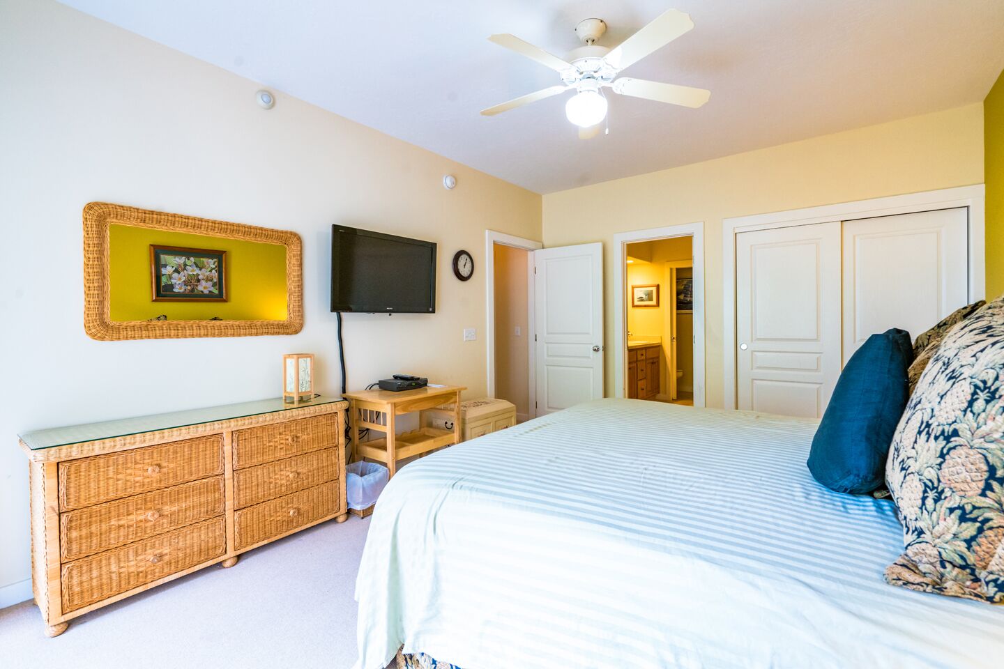 A bedroom with a bed, a television, a ceiling fan, a dresser, and a door leading to a bathroom.