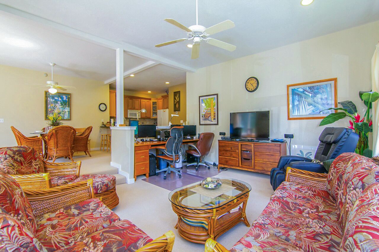 Un salon comprenant un canapé, des fauteuils, une table basse, un téléviseur et un bureau avec un ordinateur. On y trouve également un ventilateur de plafond, une horloge et une plante.